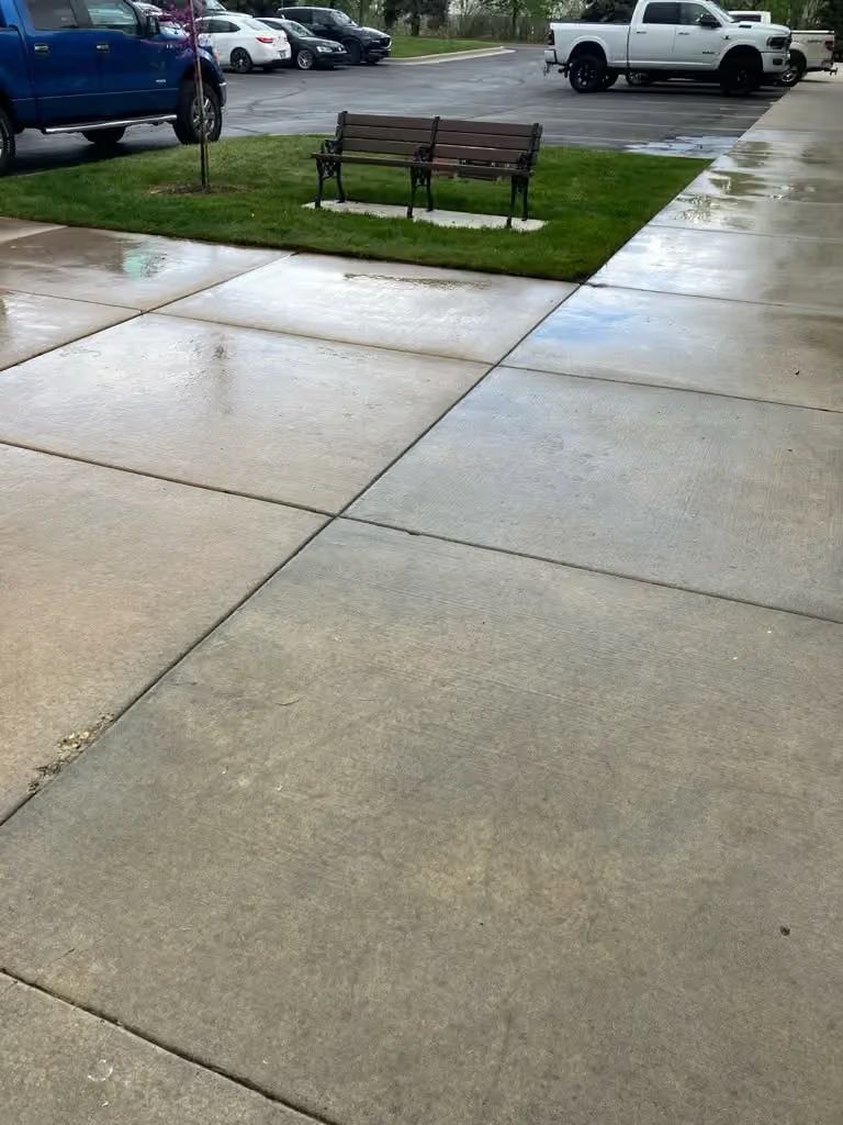 Wet concrete sidewalk beside a bench and grassy parking lot on a rainy day