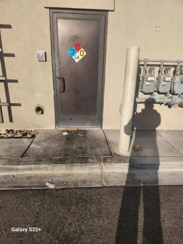 Gray utility door with hazard placard beside outdoor electrical meters and a sidewalk curb