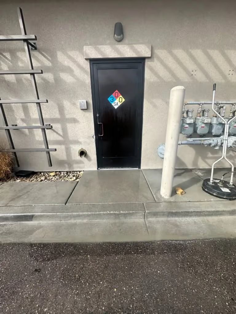 Black utility door with hazard placard on a concrete exterior wall beside gas meters and a ladder.