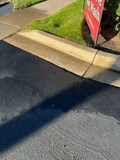 Curbside asphalt beside a concrete sidewalk with a red yard sign and grass in sunlight