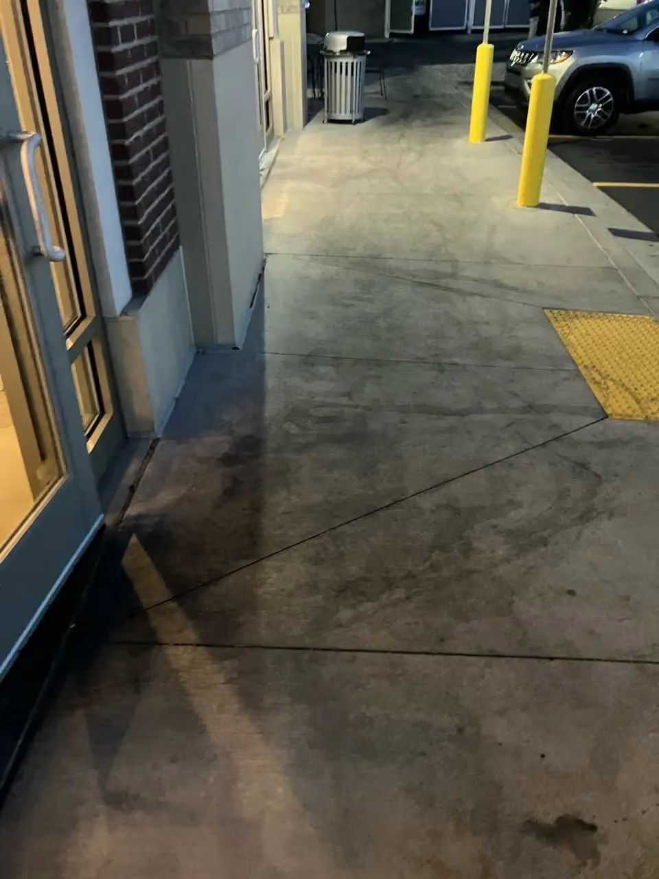 Storefront sidewalk at night with yellow bollards and parked cars.