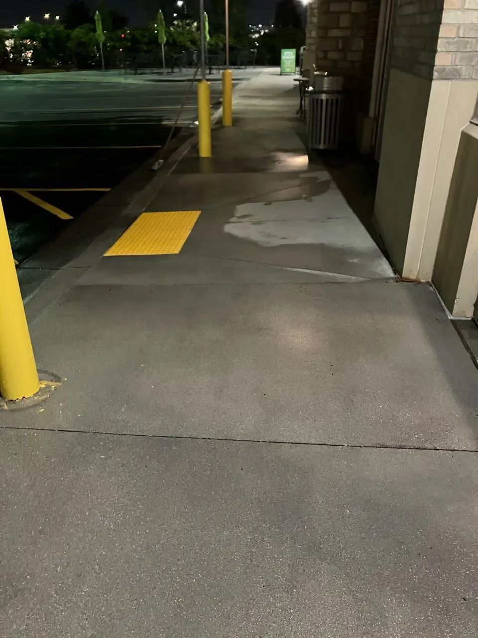 Nighttime sidewalk beside a parking lot with yellow bollards and a patched concrete ramp area