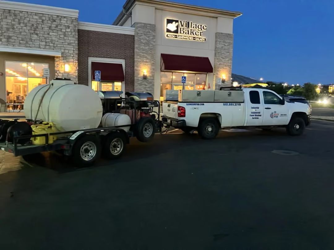 White pickup towing a small water tank trailer outside a lit storefront at dusk