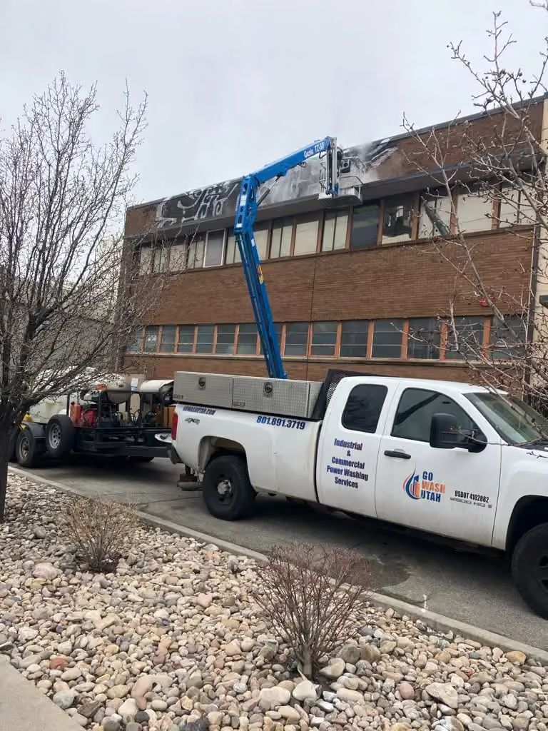 White utility truck with blue boom lift parked beside a brick building, near a gravel path and leafless trees