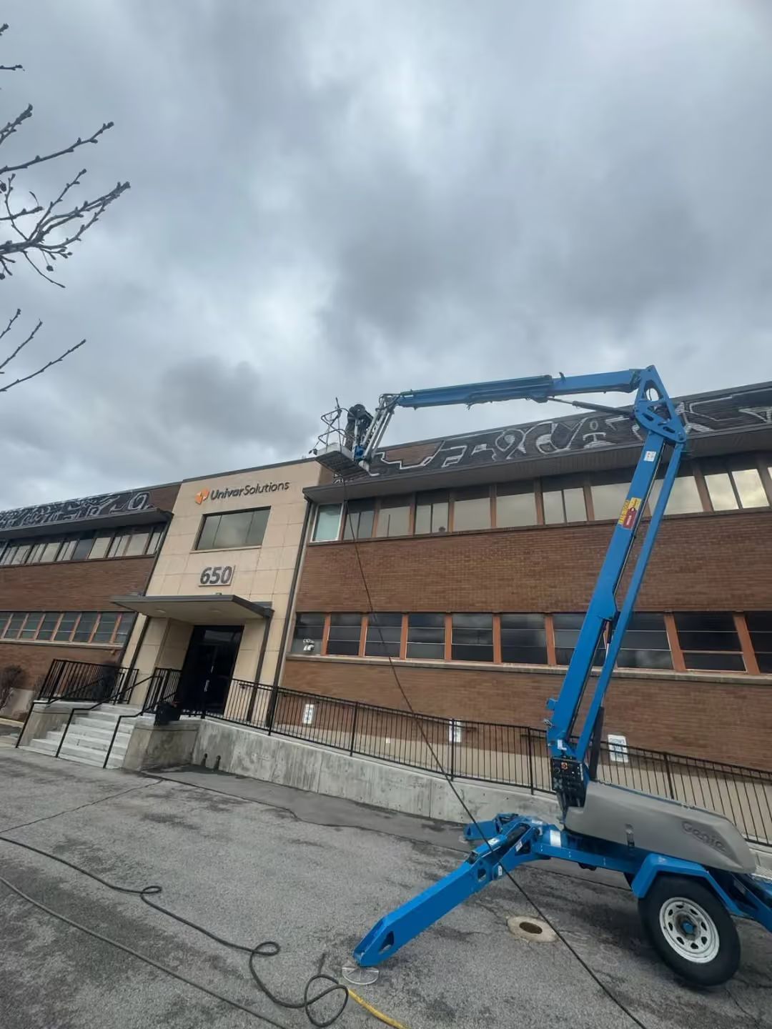 Blue boom lift beside a brick building with a damaged roof under cloudy skies