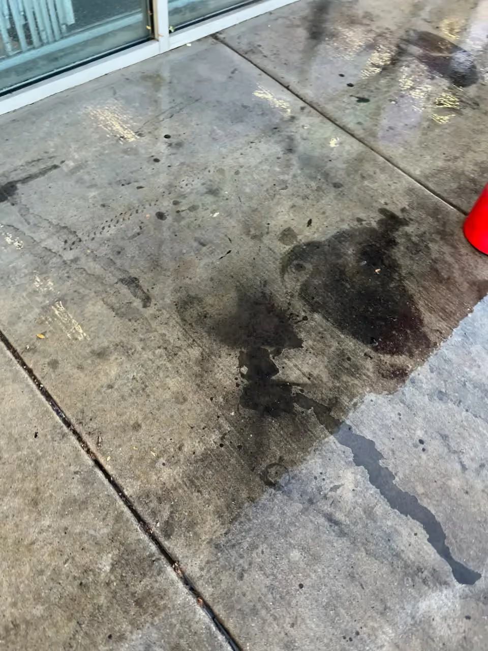 Wet concrete sidewalk with dark stains and a red bucket near a glass wall