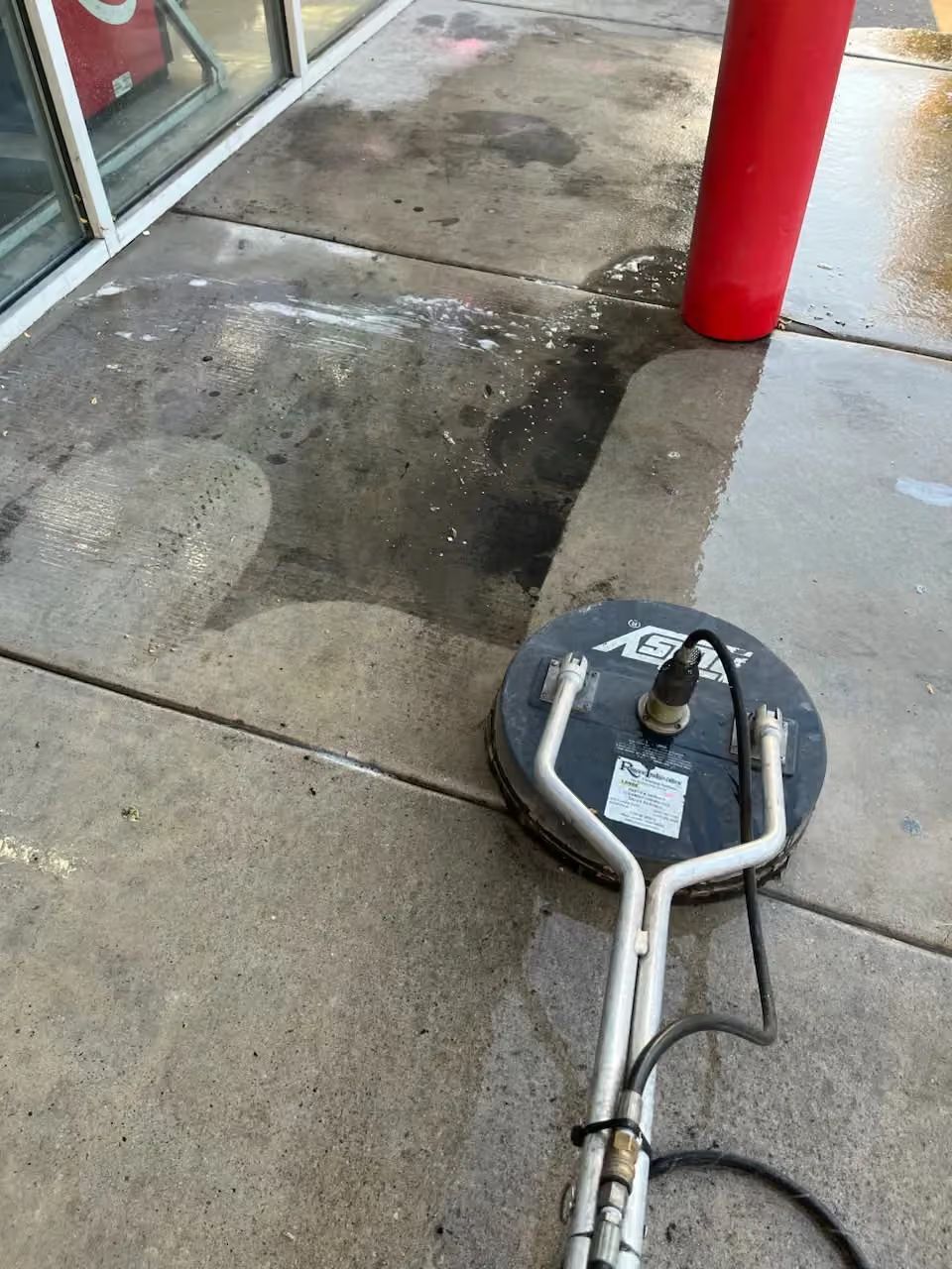 Pressure washer cleaning dirty concrete sidewalk near a red post by a storefront entrance