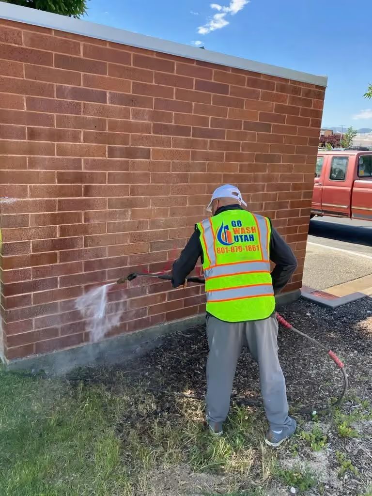Worker in neon safety vest spraying brick wall with cleaning solution outdoors.