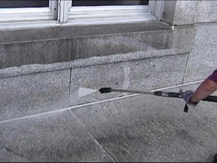 Person pressure-washing a stone building wall and sidewalk with a long spray wand.