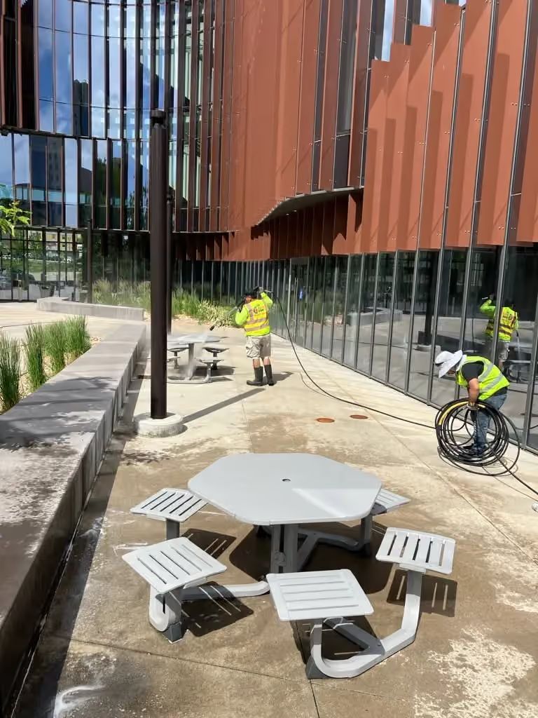 Outdoor courtyard with picnic tables and two workers in yellow safety vests near a modern building.