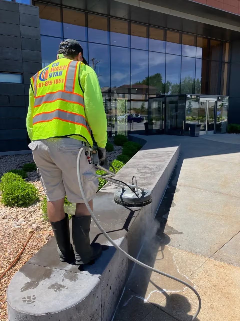 Worker in neon safety vest inspecting a cable on a concrete ledge outside a glass building.