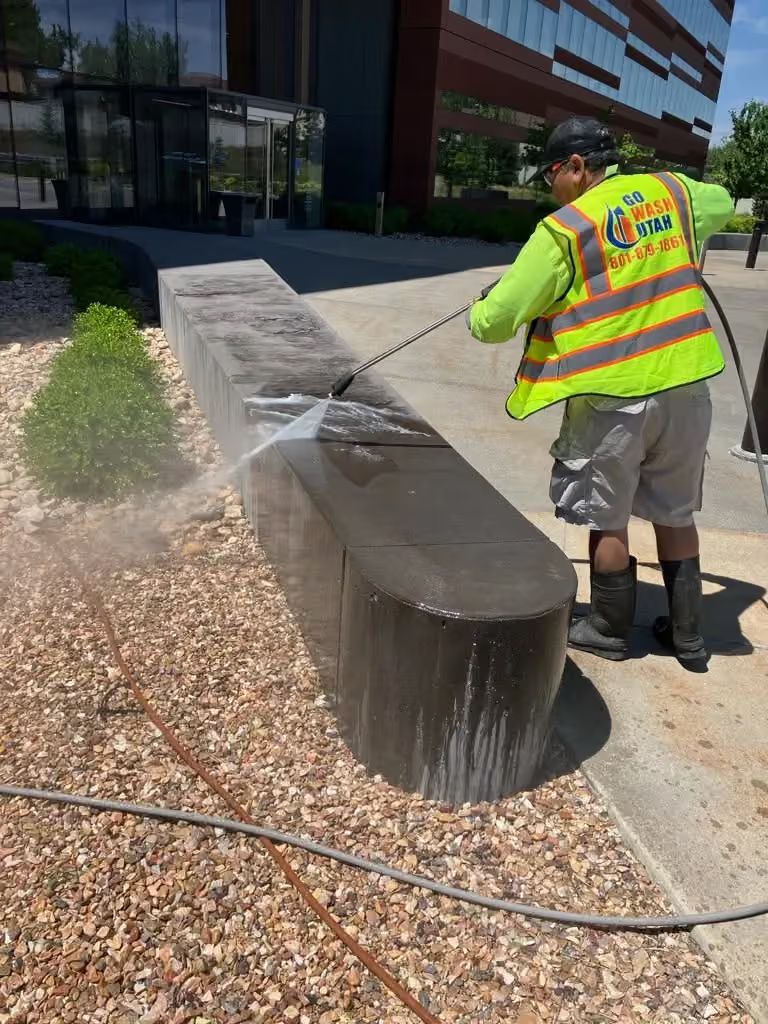 Worker spraying water on a curved concrete bench beside a modern building.