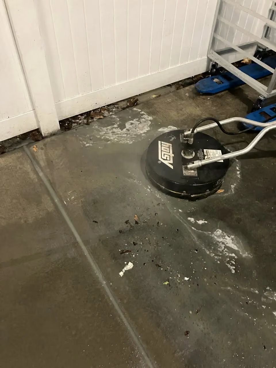 Dirty concrete floor being cleaned with a floor scrubber beside a white fence and blue ladder