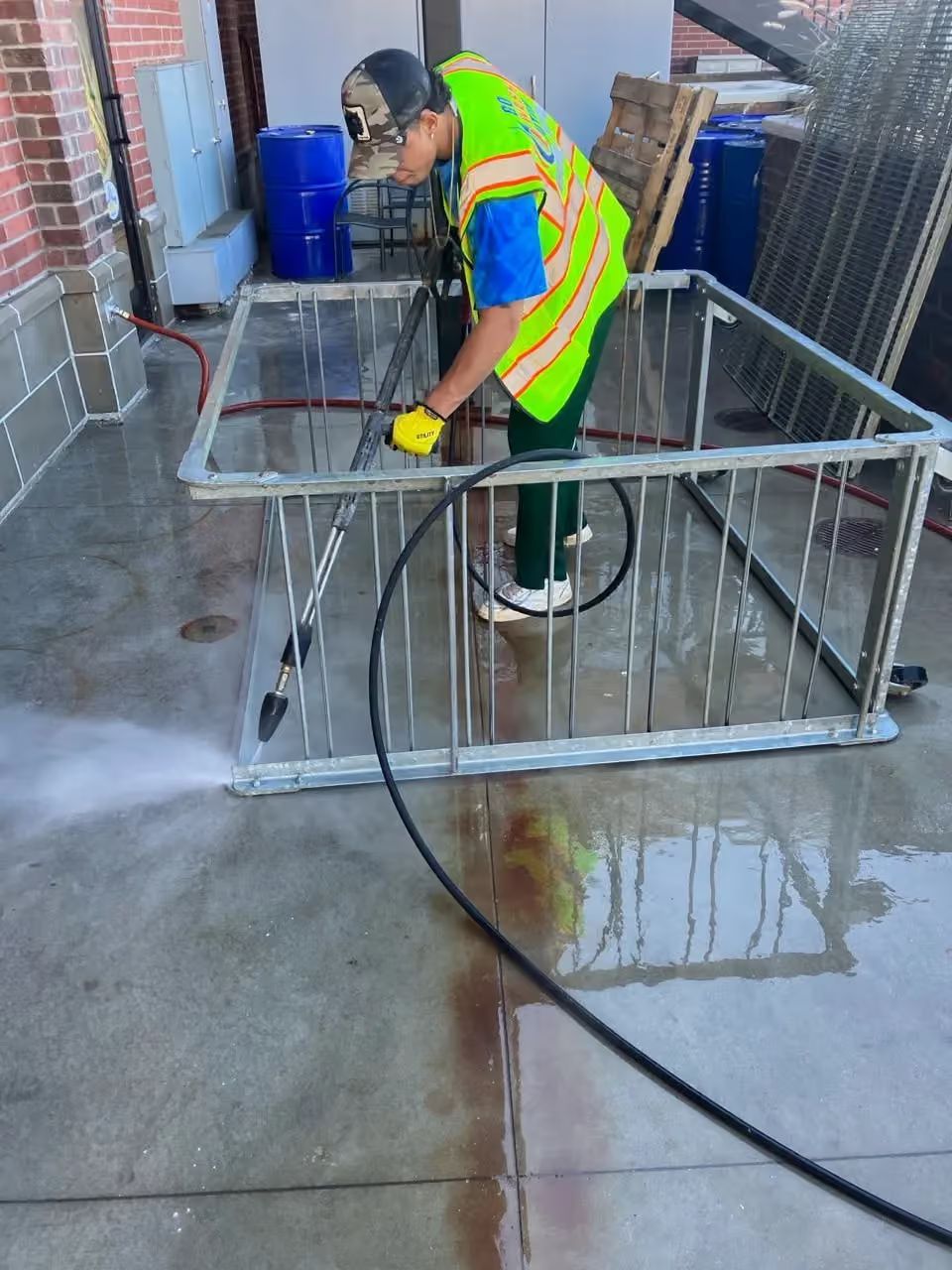 Worker in safety vest pressure-washing a metal cart in an outdoor alley.