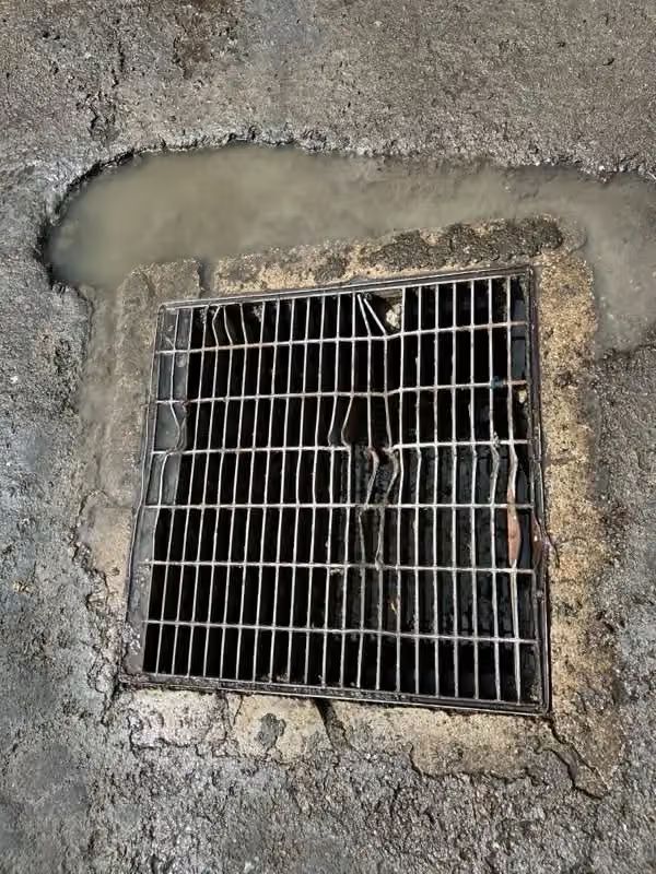 Grated drain opening in rough concrete pavement with standing water nearby