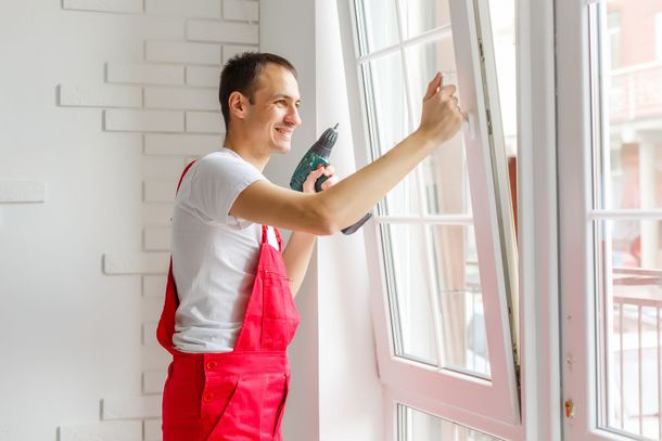 A man in red overalls is installing a window with a drill.