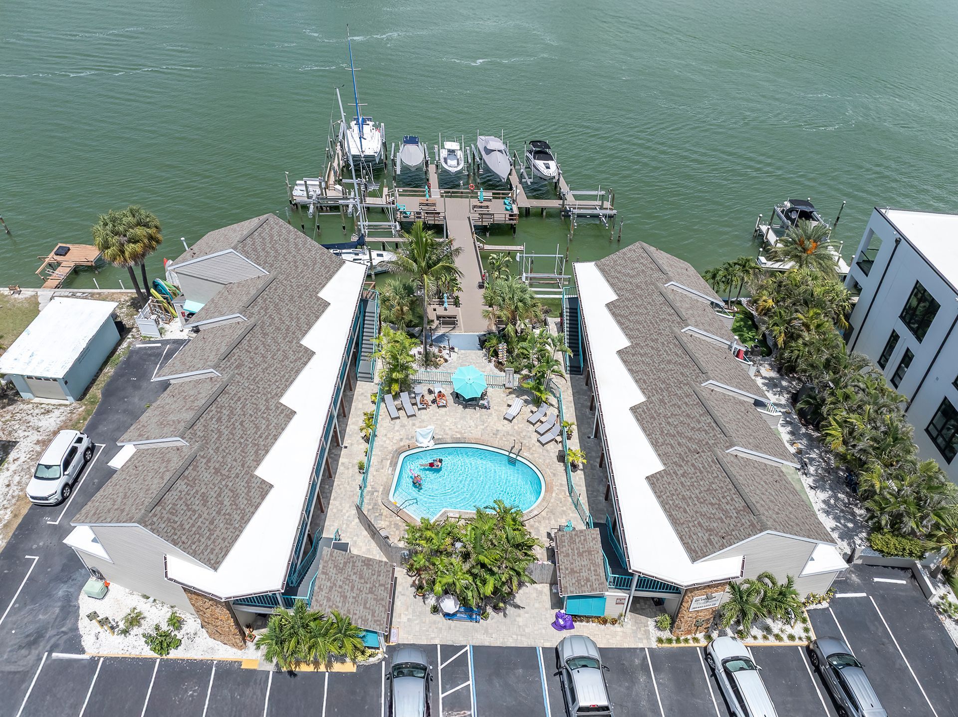 Condos with blue trim surround a courtyard with a pool, palms, and a water view.