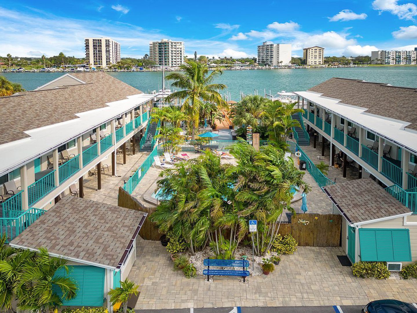 Condos with blue trim surround a courtyard with a pool, palms, and a water view.
