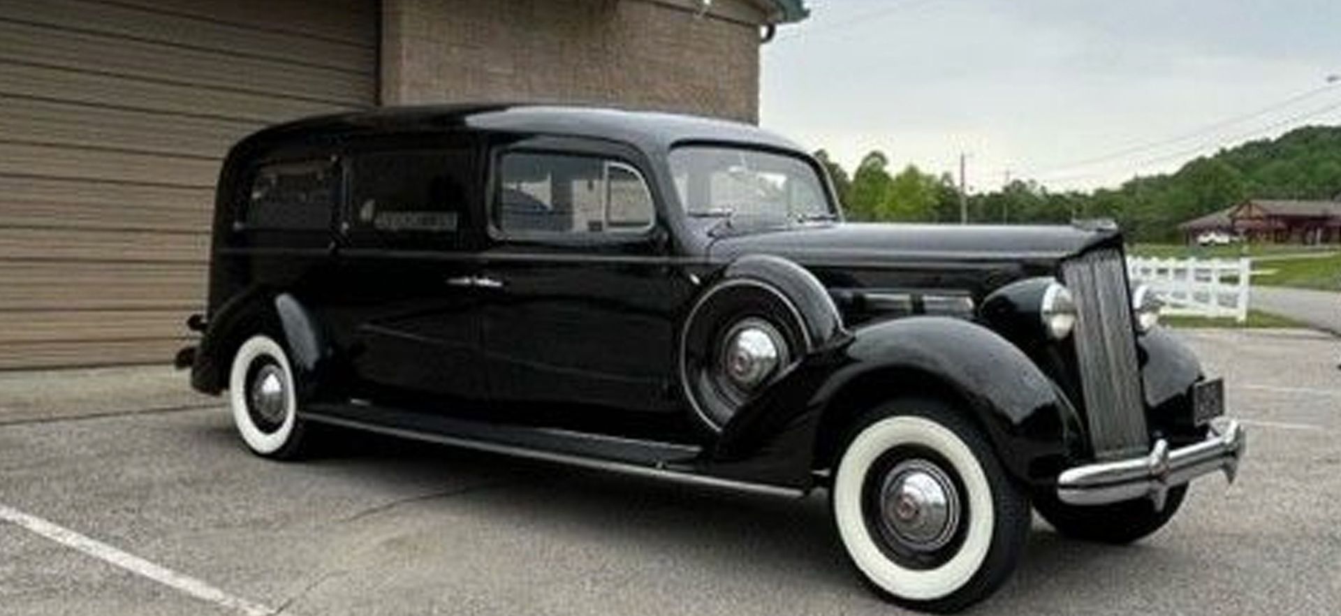 An old black car with white tires is parked in front of a garage.