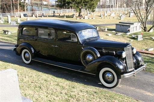 An old black car with white tires is parked in a cemetery.
