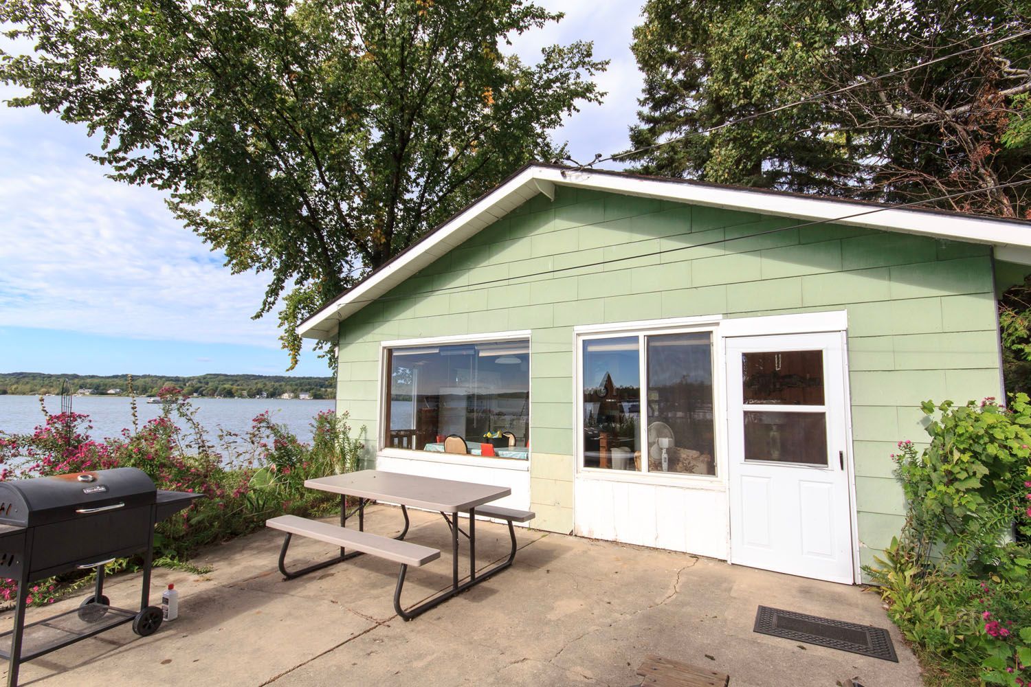 A light green cottage with white trim stands by a lake, featuring a patio with a picnic table and a charcoal grill.