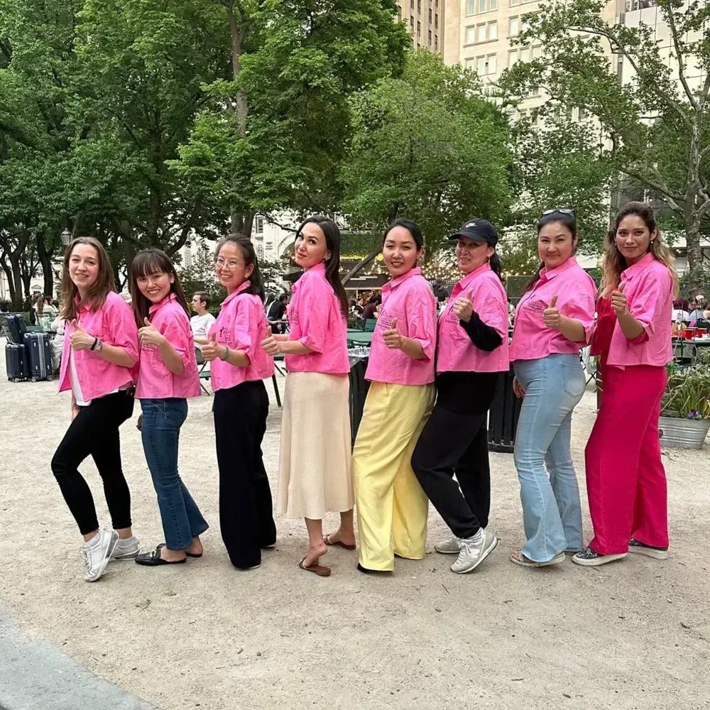 Eight people wearing pink tops stand in a row outdoors in a park, posing and smiling for the camera.