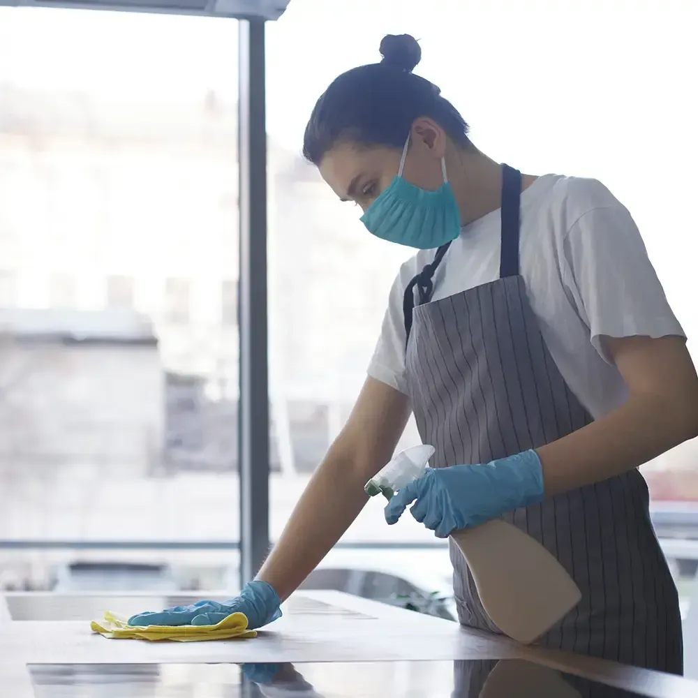 A person in a mask, gloves, and apron cleaning a surface with a spray bottle and yellow cloth near a window.