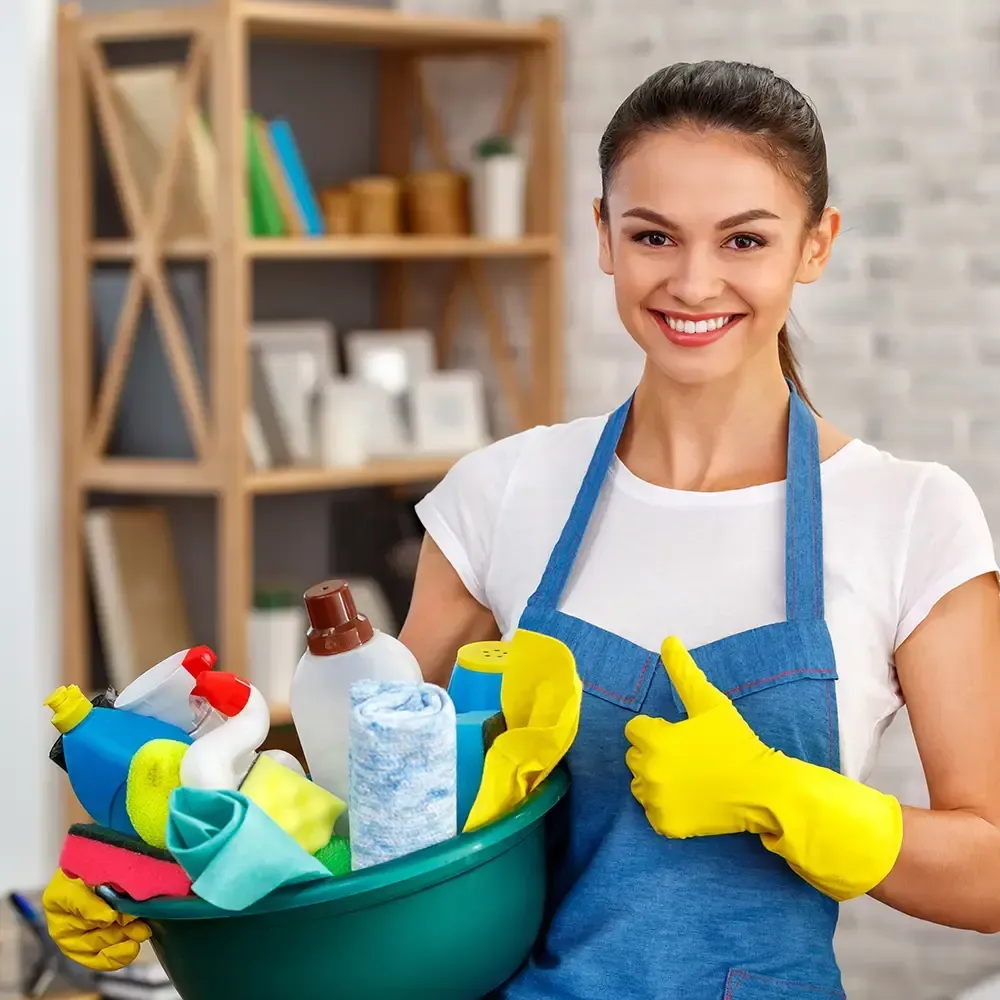 A smiling person in a blue apron and yellow gloves holds a basin filled with cleaning supplies and cloths.
