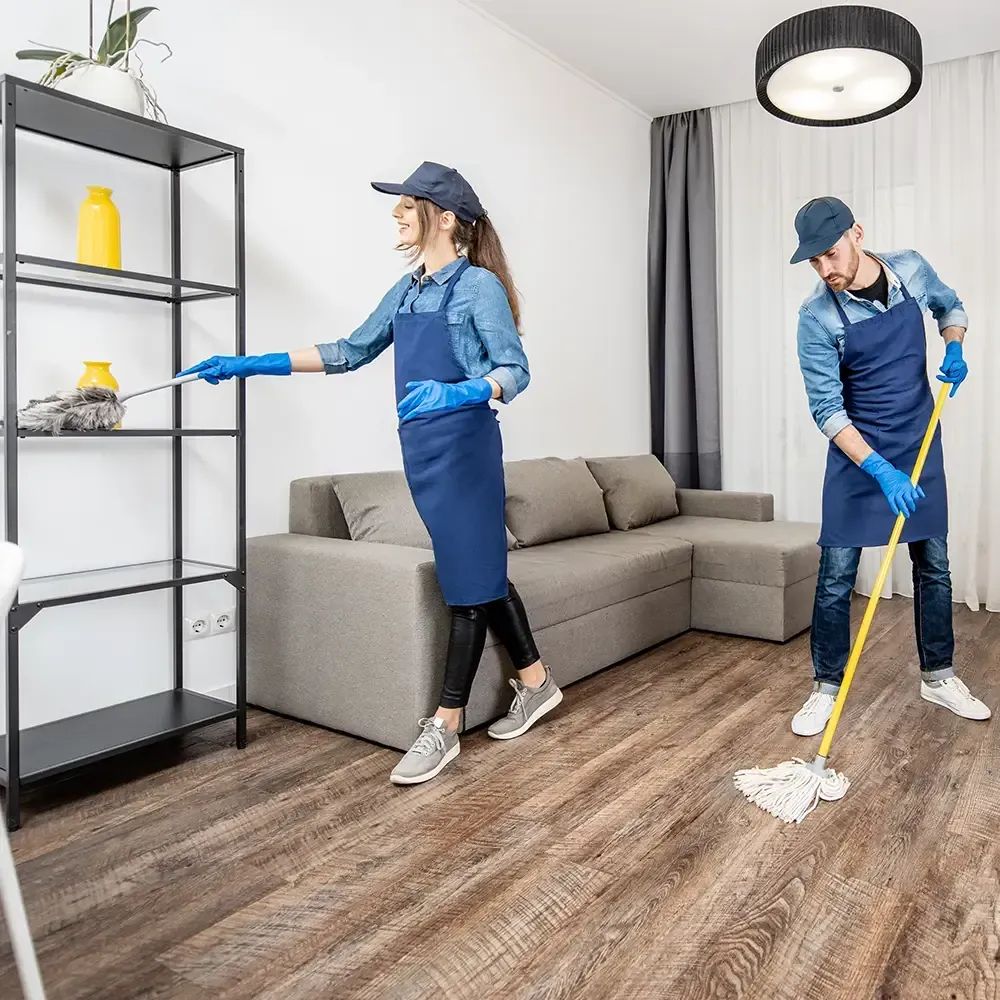 Two people in cleaning uniforms are working in a modern living room; one dusts a metal shelf, the other mops the floor.