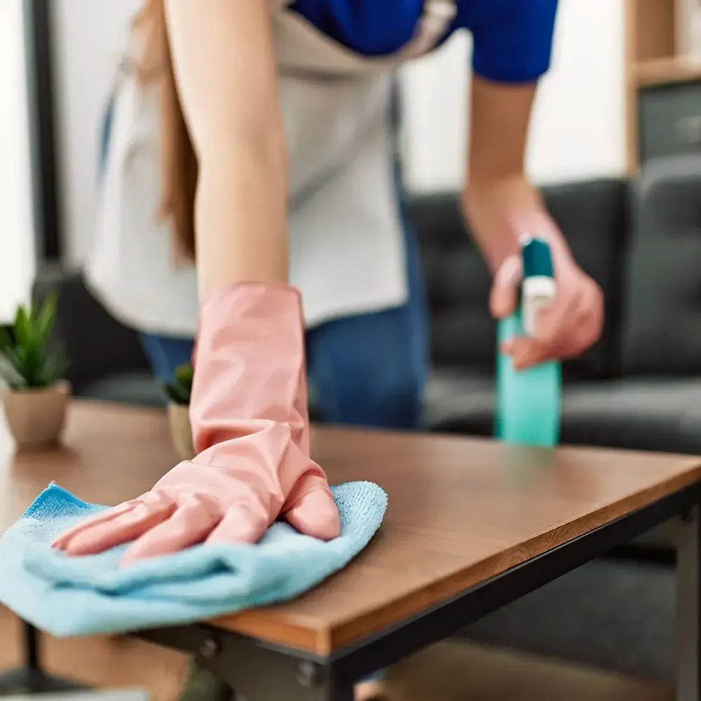 A person wearing a blue shirt, apron, and pink rubber gloves cleans a wooden table with a blue cloth and spray bottle.