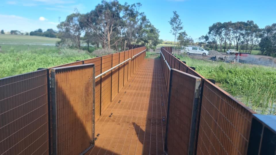 A wooden walkway leading to a field with trees in the background.
