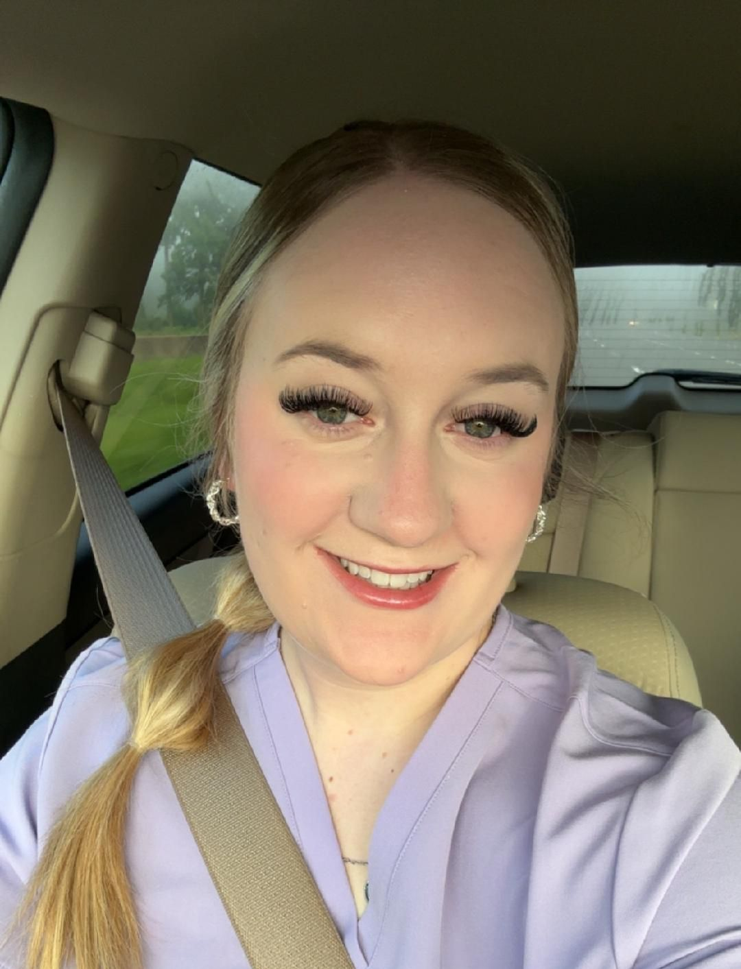 Woman in car, wearing lavender scrubs, smiles at camera. Blonde hair in braid, long lashes, silver earrings.