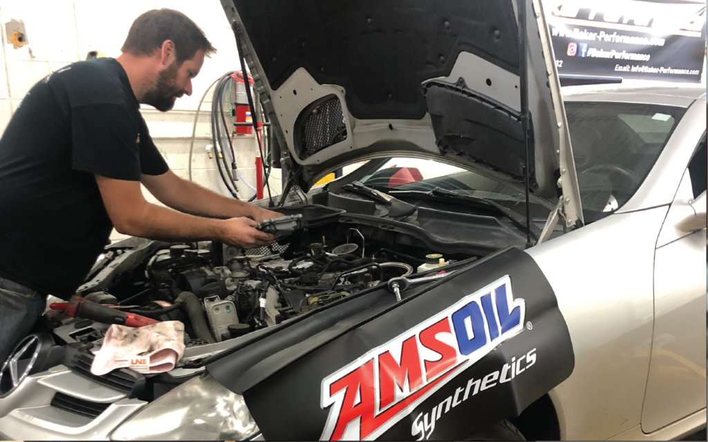 Mechanic working on a car engine, under the hood, in a garage. An AMSOIL cover protects the car. | SB Automotive