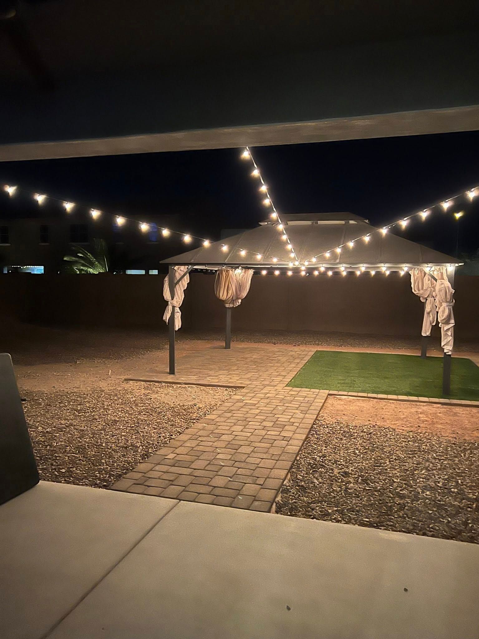 String lights over a backyard patio at night, with a stone path and small grass area.