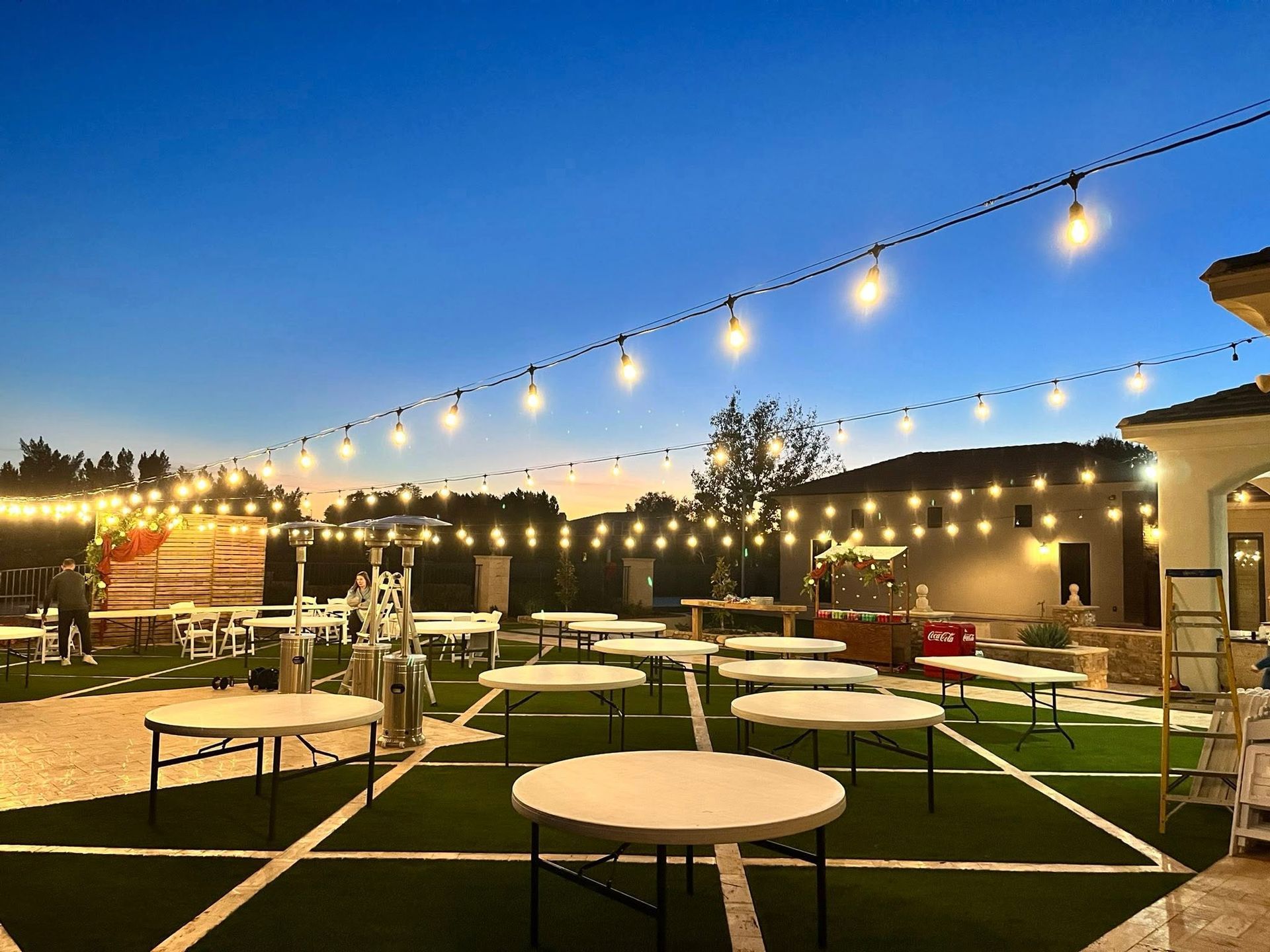 Outdoor patio seating area at dusk with string lights, round tables, and artificial grass