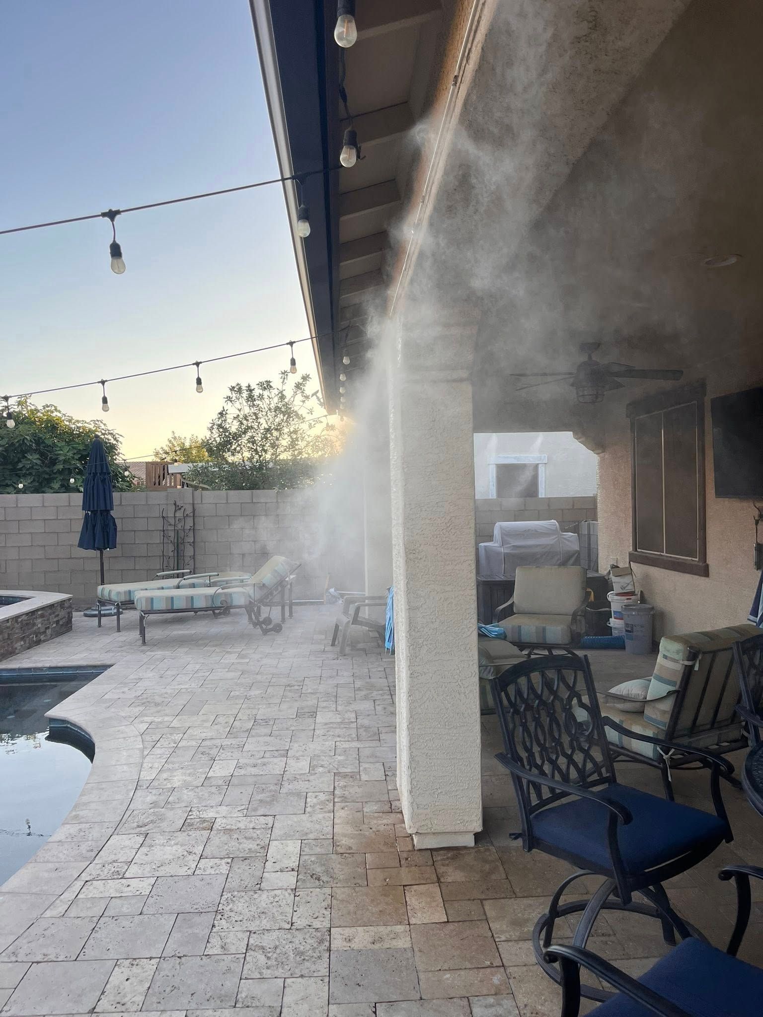 Backyard patio with a misting spray under the roof overhang at dusk, string lights and outdoor seating visible