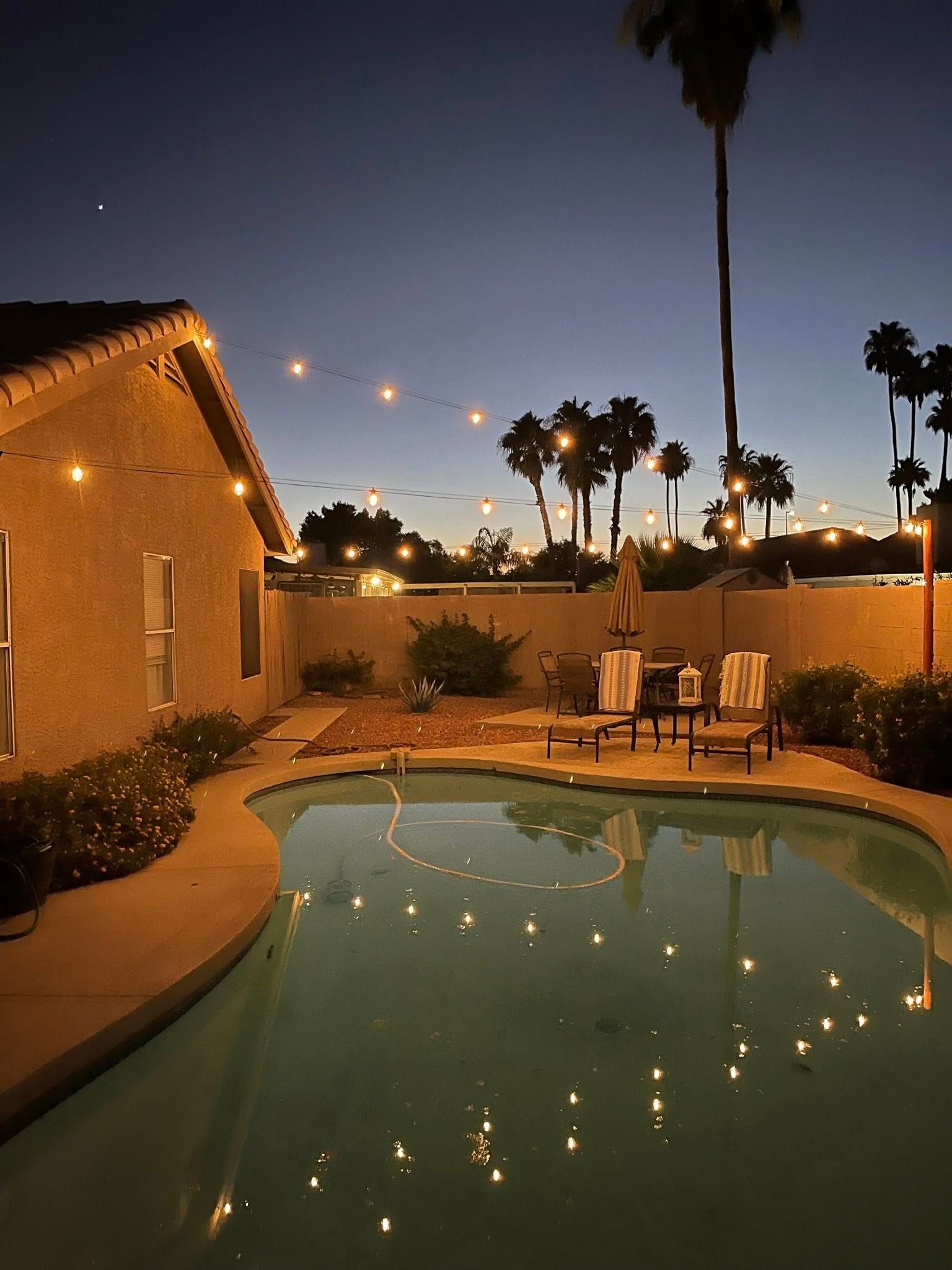 Backyard pool at dusk with string lights reflecting on the water beside a house and patio chairs