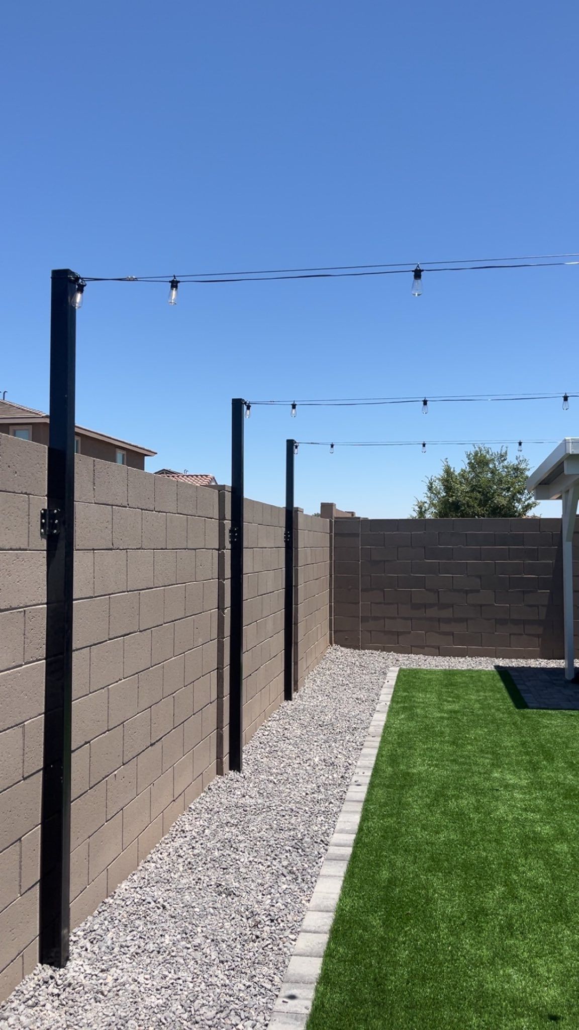 Narrow backyard walkway with gravel, black posts, tan block walls, string lights, and green grass under blue sky
