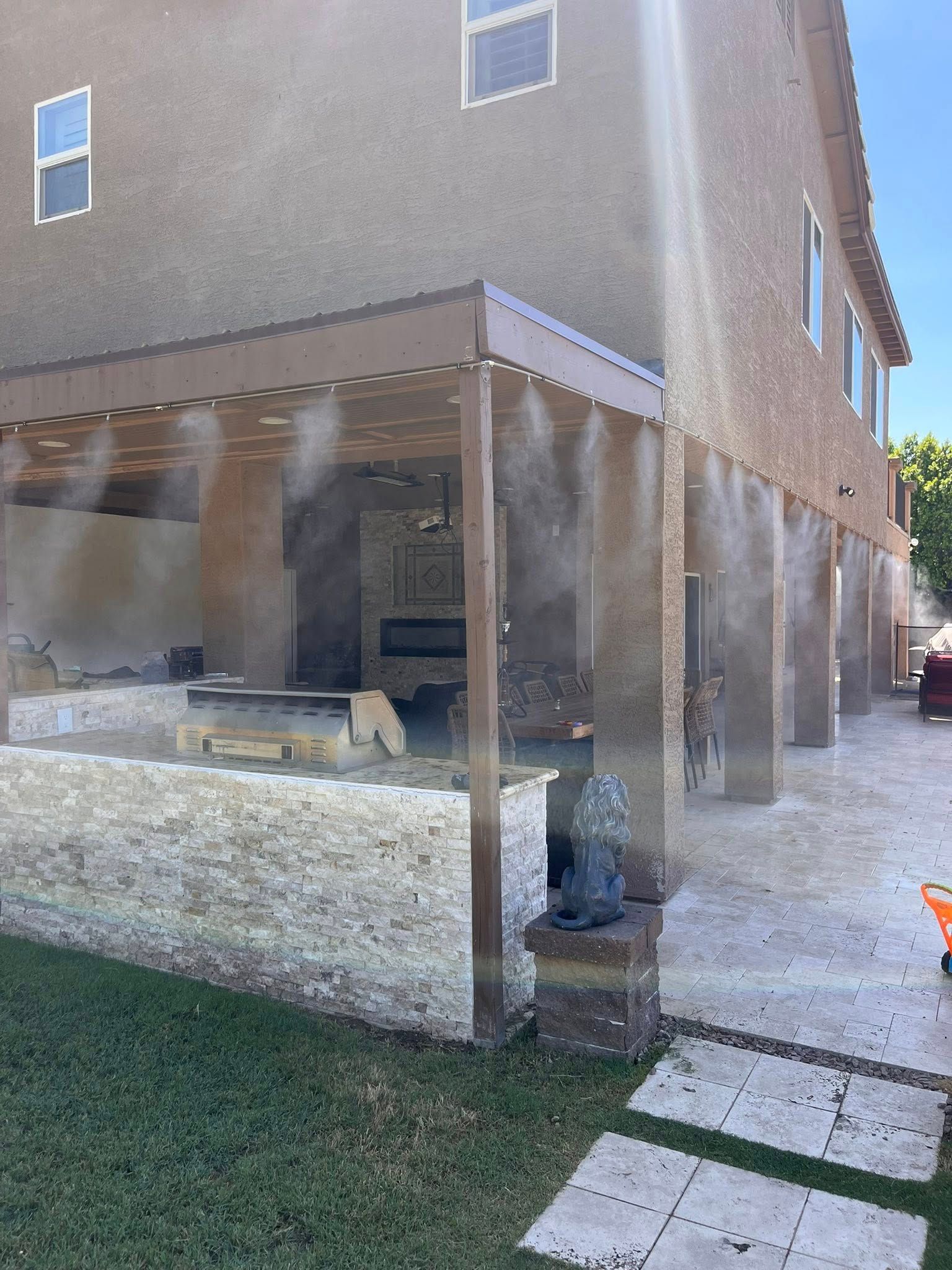 Covered patio beside a tan house, with a gravel yard and stepping stones in sunlight.