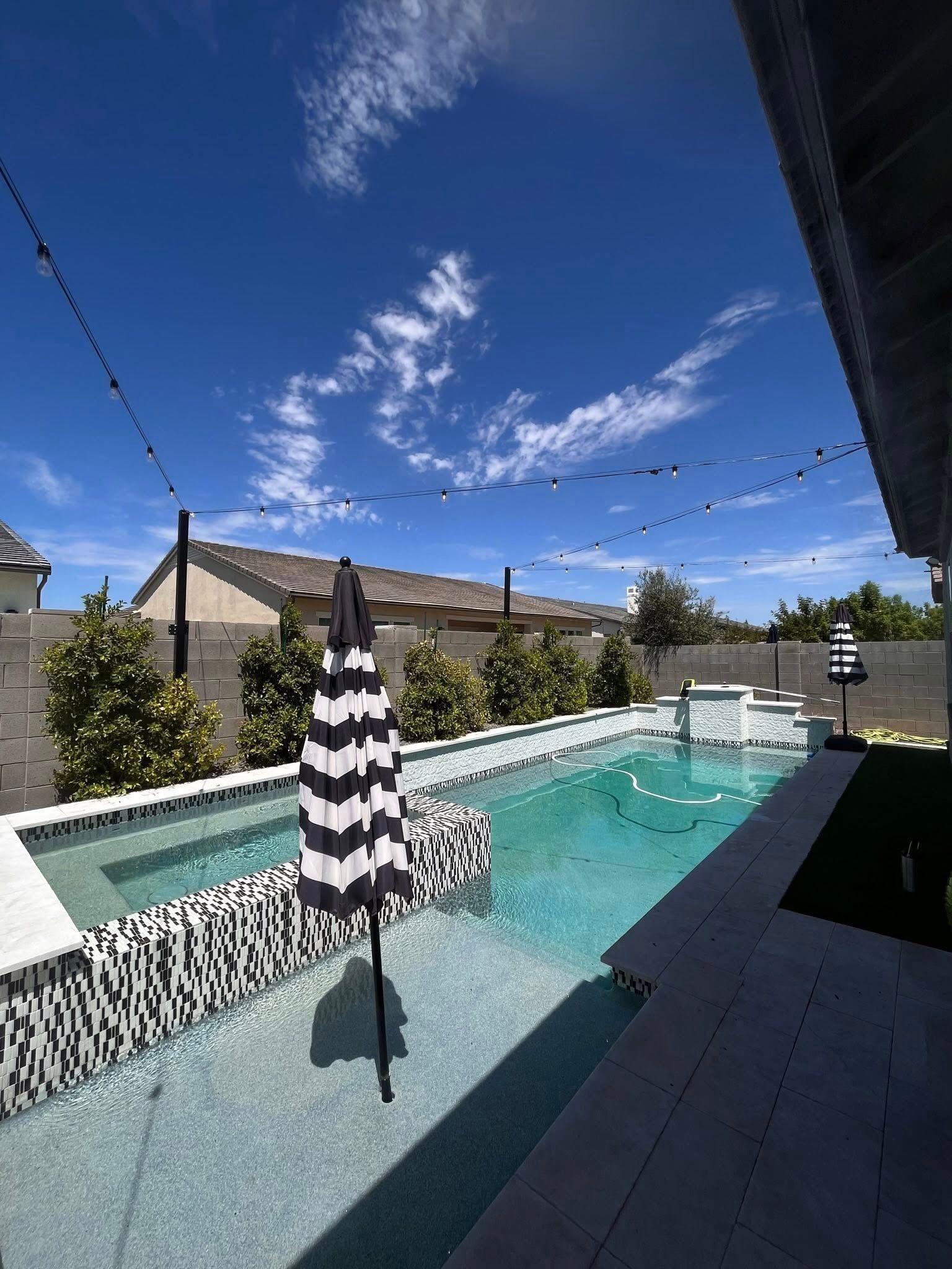 Modern backyard pool with turquoise water, striped umbrella, and string lights under a clear blue sky