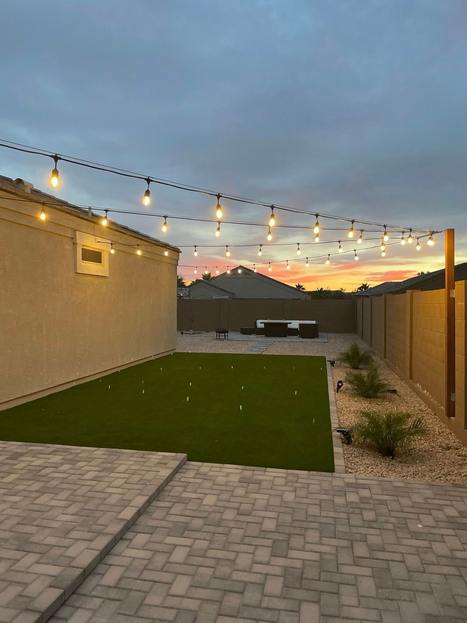 Backyard patio at dusk with string lights, artificial turf, and a sunset sky