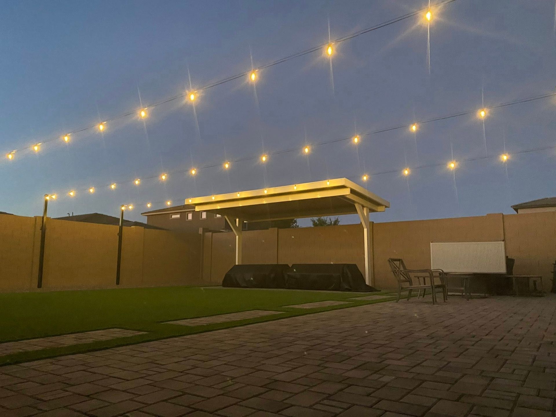 Backyard patio at dusk with string lights, a wooden pergola, brick paving, grass, and outdoor chairs and table.