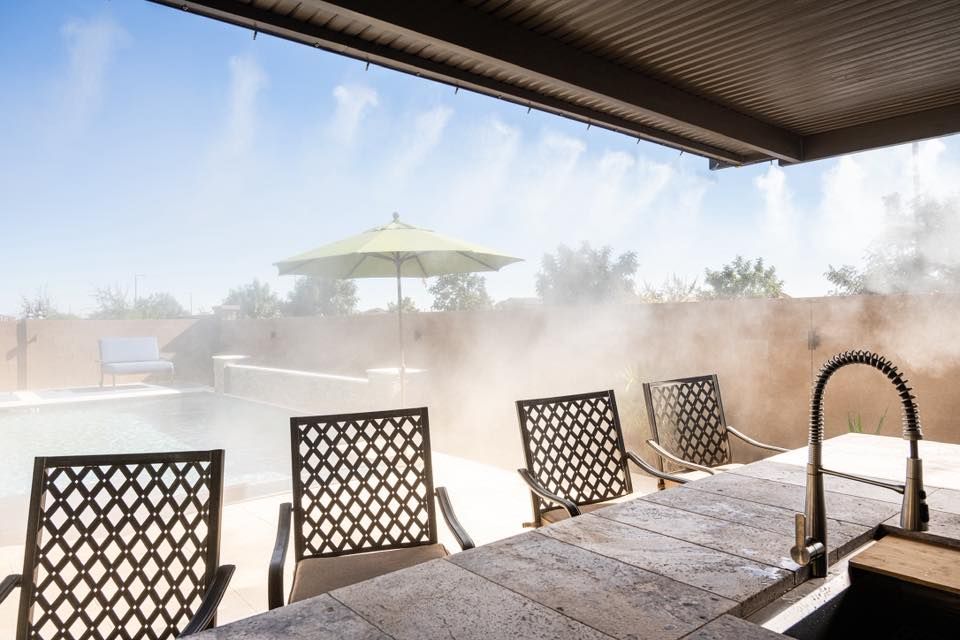 Patio chairs beside a table and outdoor sink, looking out onto a misty backyard pool area