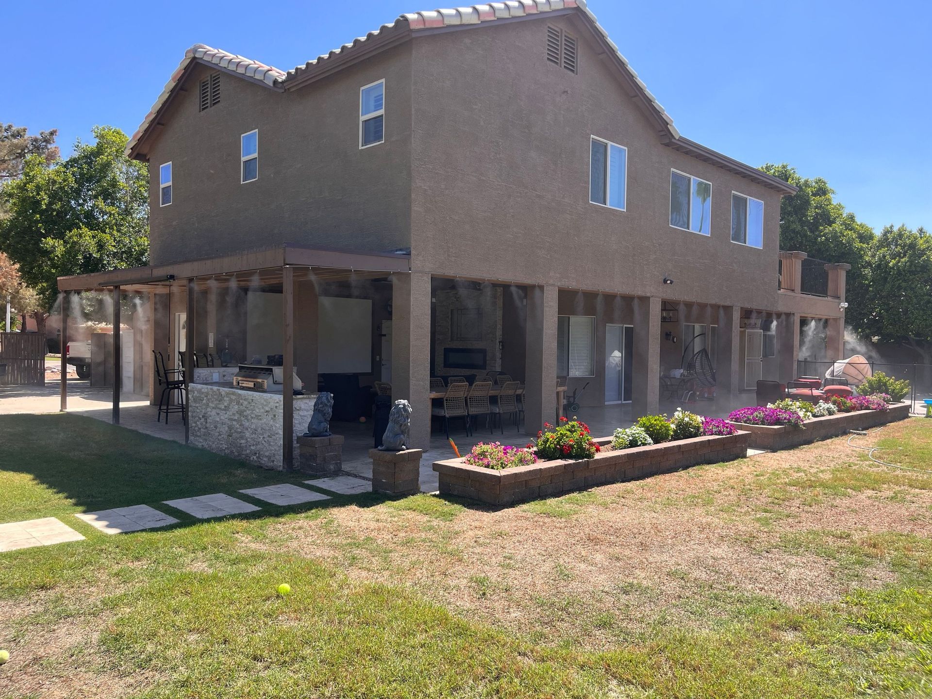 Two-story tan house with a covered patio, flower beds, and a grassy yard under a clear blue sky