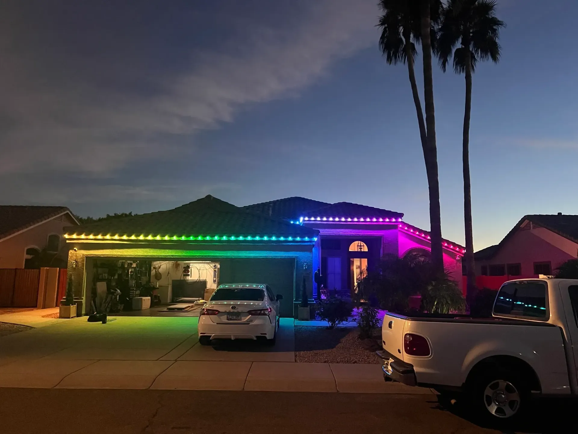 Single-story house at dusk with rainbow LED roof lights, parked cars, and tall palm trees.