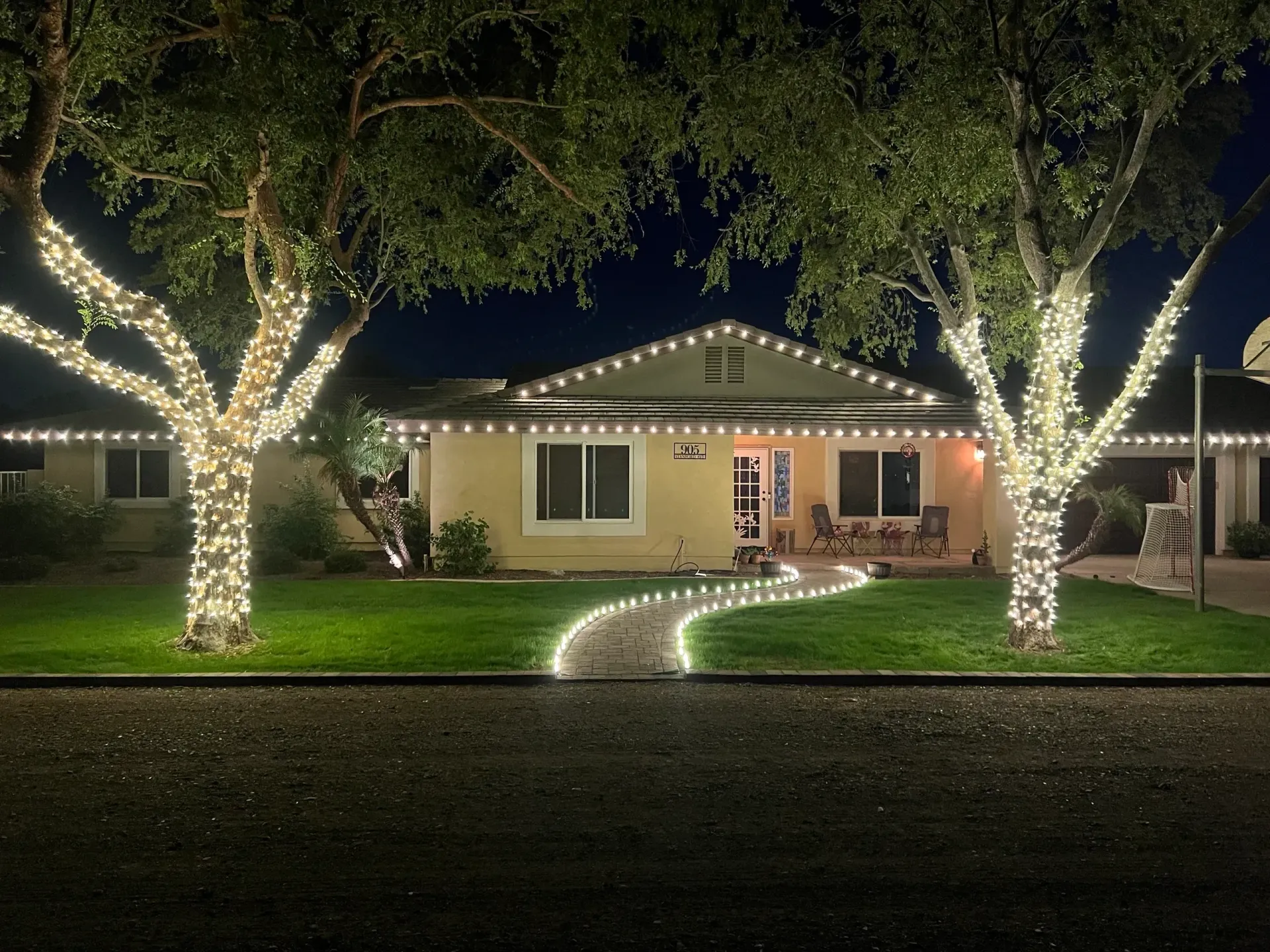 Single-story house at night with trees wrapped in white lights and a lit walkway to the front door.