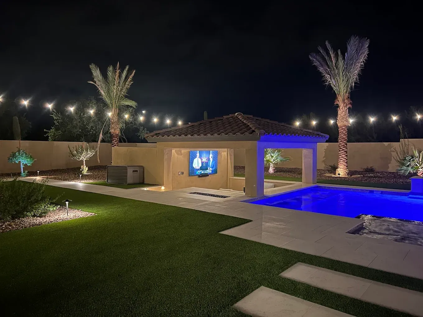 Modern backyard at night with lit pool, palm trees, lawn, and house patio lights