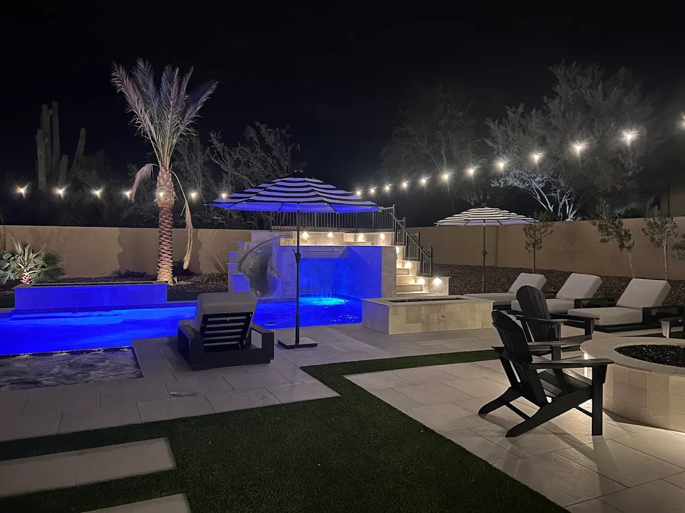 Nighttime backyard with lit blue pool, waterfall, lounge chairs, and string lights around a patio.