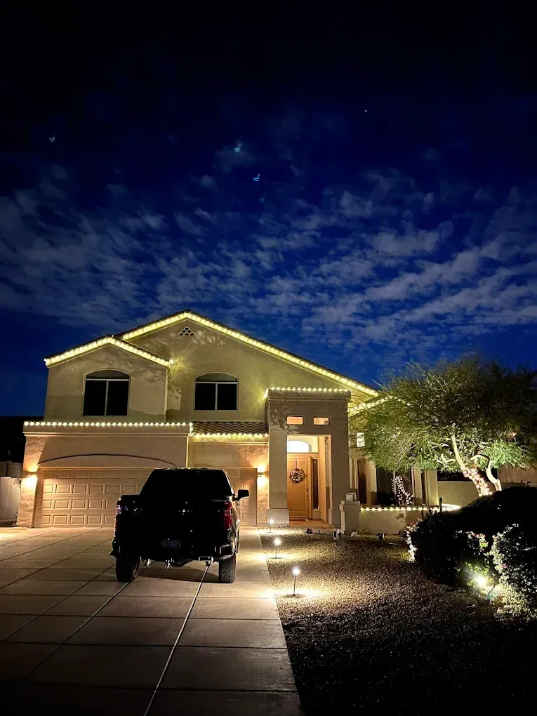 Nighttime view of a lit two-story house with cars in the driveway under a cloudy blue sky