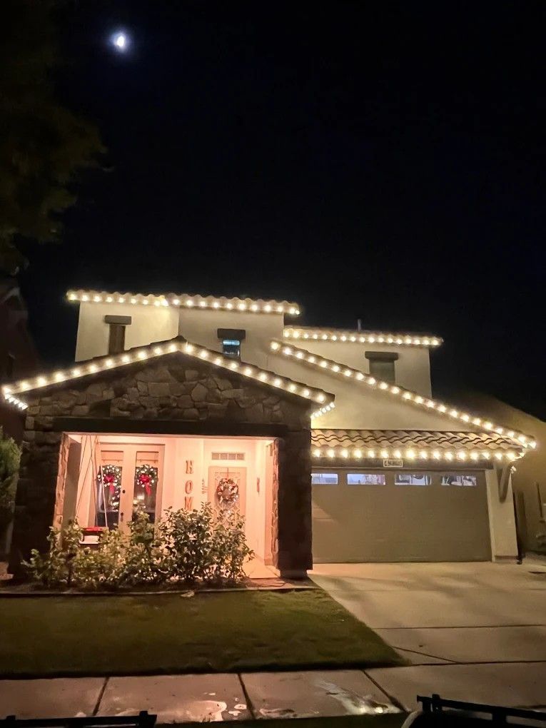 House at night outlined with white string lights, warm porch glow, and a lit garage door.
